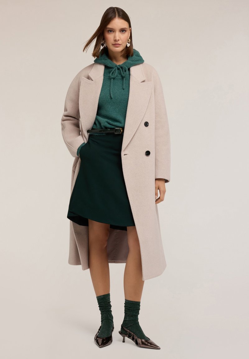 Abrigo largo de color rosa claro con botones negros sobre una sudadera verde, combinado con una falda verde oscura, calcetas texturizadas verdes y tacones negros puntiagudos.