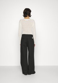 Witte gebreide top met lange mouwen en ribbelstructuur, gecombineerd met losse zwarte broek met achterzakken en een label op de tailleband.