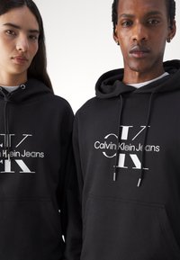 Crne dukserice imaju veliki bijeli logo s tekstom "Calvin Klein Jeans" i "CK". Materijal izgleda meko, s vezicama i prednjim džepom.