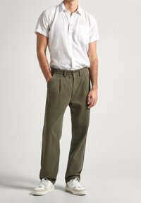 Camicia bianca in lino a maniche corte con taschino, abbinata a pantaloni plissettati verdi oliva e sneakers bianche su uno sfondo neutro.