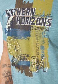 Turquoise graphic t-shirt featuring abstract patterns in blue, green, and brown, with "NORTHERN HORIZONS" text and number "84." Short sleeves.