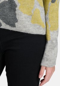 Sweter dziergany w szarym kolorze z abstrakcyjnymi wzorami w żółtym i czarnym. Miękka faktura, długie rękawy i ściągacze. W zestawie z czarnymi spodniami.