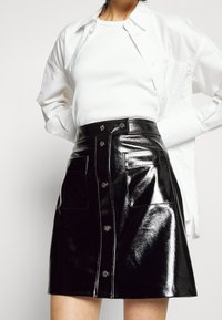 Mini-jupe en cuir verni noir avec une taille ajustée, une texture brillante, une fermeture à boutons à l'avant et deux poches latérales, associée à un haut blanc et une chemise.