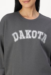Sudadera gris con cuello redondo. Presenta "DAKOTA" en letras blancas en arco a través del pecho. Textura suave, diseño casual.