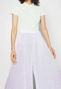 Vihreä polo-paita, jossa on kaulus ja nappikiinnitys, yhdistettynä valkoisiin laskostettuihin leveälahkeisiin housuihin, joissa on vaaleanvioletteja pystysuoria raitoja.