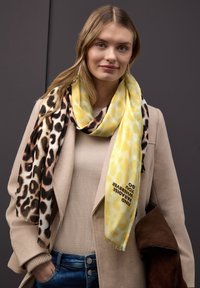 Jeune femme portant un manteau beige et une écharpe imprimée léopard jaune et marron avec du texte, tenant une veste en suède marron sur un fond sombre.