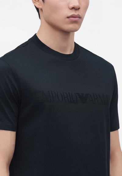 Νέος άνδρας που φοράει μαύρο T-shirt Emporio Armani με διακριτικό μαύρο λογότυπο στο στήθος, στέκεται μπροστά σε απλό φόντο.