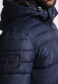 Chaqueta tipo puffer en color navy con textura acolchada, que incluye una capucha forrada en marrón, cordones y un parche en la manga izquierda. Diseño casual.