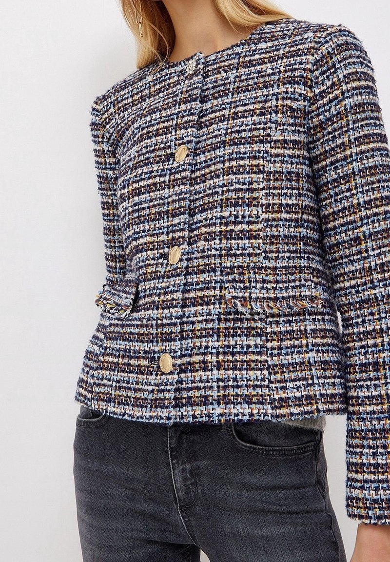 Giacca in tweed con un mix di fili blu, marroni e gialli, bottoni dorati e due tasche frontali con dettaglio frangiato all'orlo.