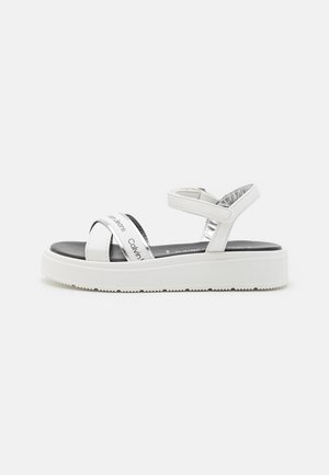 White platform sandals with cross-strap design, featuring glossy silver accents and a textured sole. Black footbed with brand text.