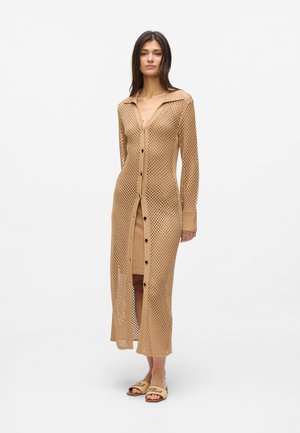 Žena v dlhom, béžovom otvorenom pletenom kardigáne na gombíky, oblečená na béžové šaty a béžové šľapky, stojaca pred jednofarebným bielym pozadím.