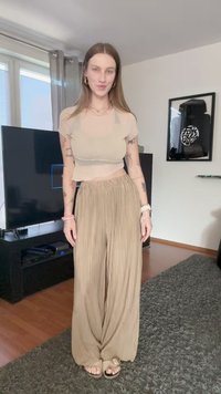Beige lühike topp, millel on lainelised ääred, koos kõrge vöökoha ja volditud beežide pükste kombinatsiooniga. Kanda kergete sandaalide paaris. Heledates toonides ruumis.