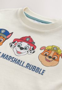 Cream sweatshirt featuring cartoon dog characters with vibrant colors: red and yellow hats, black spots, and prominent facial features. Text below characters.