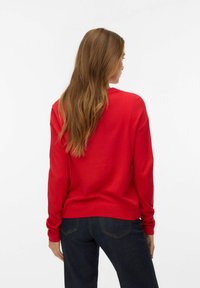 Maglione rosso lavorato a maglia con vestibilità comoda, polsini e orlo a coste. Il modello indossa jeans scuri. Capelli che ricadono sulla schiena. Sfondo bianco semplice.