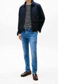 Hombre que lleva una chaqueta azul marino, una camisa a rayas en blanco y negro, jeans azules y mocasines de cuero marrón oscuro, de pie contra un fondo blanco.