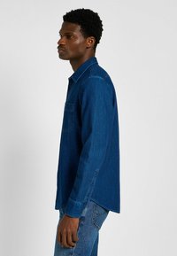 Niebieska koszula dżinsowa z długimi rękawami, zapięciem na guziki, jedną kieszenią na piersi oraz kontrastującym szwem. Teksturowana tkanina i luźny krój.