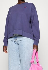 Persona che indossa un felpa viola di Levi's e jeans blu chiaro, mentre tiene una borsa rosa pallido per i manici.