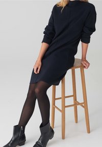 Femme portant une robe noire à manches longues, des collants noirs et des bottines noires, assise sur un tabouret de bar en bois sur un fond gris.