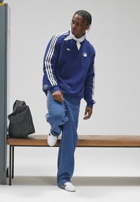 Marineblå genser med hvite striper, avslappede lyseblå jeans og hvite sneakers. Stylet med et hvitt bandana, sittende på en trebenk.