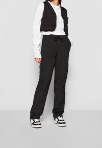 Svarta cargo-byxor med dragsko i midjan, parat med en svart ärmlös väst med flera frampocket. Bärs med svart och vit sneakers.