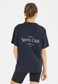 Tummansininen t-paita, jossa on valkoinen "Sports Club" -grafiikka selkäpuolella; lyhythihaiset, rento malli, valmistettu pehmeästä puuvillakankaasta.