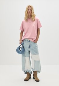 Kvinde med løst blont hår iført en pink grafisk t-shirt, patchwork vidde ben jeans, brune støvler og holder en lille denim håndtaske.