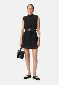 Abito nero a trama con collo alto, vita cintrata da una ampia cintura nera. Gli accessori includono una piccola borsa nera e scarpe flat.