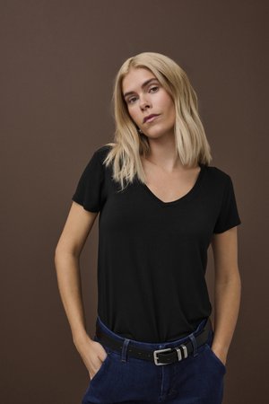 Blondínka s ramenami dlhými vlasmi v čiernom tričku s výstrihom do V a modrých džínsoch s čiernym opaskom, stojaca s rukami vreckách na pozadí hnedej steny.