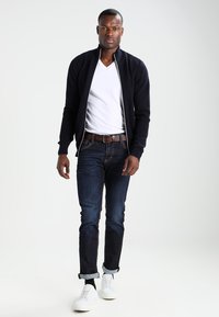 Marineblå, quiltet bomberjakke over en hvit V-hals t-skjorte, kombinert med mørkeblå jeans og hvite sneakers, med oppbrettede mansjetter på jeansene.