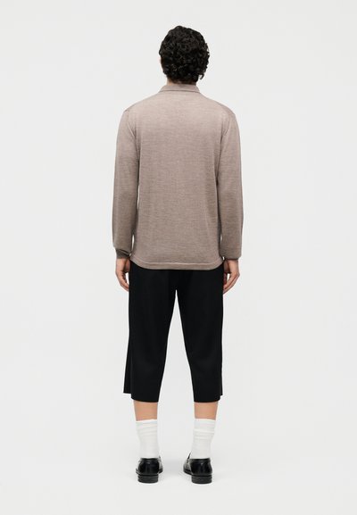 Άνδρας με σγουρά μαλλιά στέκεται με την πλάτη γυρισμένη, φορώντας μπεζ πουκάμισο με μακριά μανίκια, μαύρο παντελόνι μέχρι τον αστράγαλο, λευκές κάλτσες και μαύρα παπούτσια, σε απλό φόντο.