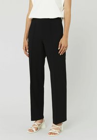 Pantalons noirs taille haute à coupe droite, en tissu texturé avec des plis à l'avant, associés à des sandales à talons carrés blanches avec des lanières.