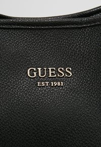 Surface en cuir noir texturé avec logo en métal argenté « GUESS EST 1981 » centré dessus.