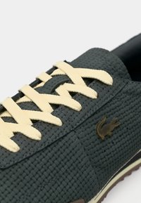 Σκούρο πράσινο υφασμάτινο αθλητικό παπούτσι με μπεζ κορδόνια και μικρό μεταλλικό λογότυπο κροκόδειλου στο πλευρικό πάνελ.