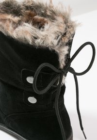 Bota de ante negra con forro de pelaje sintético, punta redonda y botones laterales. Cuenta con un lazo negro atado en la parte delantera para un ajuste seguro.