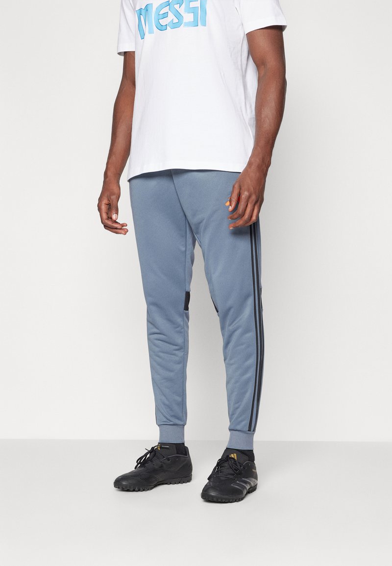 Lysblå træningsbukser med sorte sidelister og et tapered design. Båret med sorte sportssko og en hvid t-shirt med blå tekst.