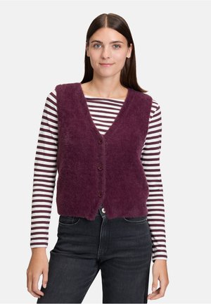 Giovane donna con capelli castani lisci che indossa un gilet viola peloso con bottoni sopra una maglietta a maniche lunghe bianca e viola a righe e jeans scuri.