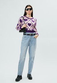 Lila und rosa gemusterter Pullover mit Herzmotiven, kombiniert mit hellblauen Jeans und schwarzen klobigen Schuhen. Mit einer schwarzen Tasche accessoriert.