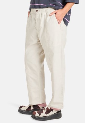 Beige trousers with an elastic waistband, side pockets, and a loose fit. Worn with patterned clogs featuring a cow print design.