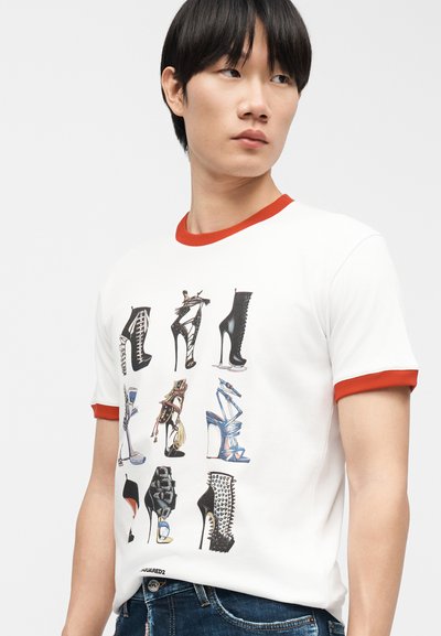 Νεαρός άντρας με μαύρα μαλλιά που φοράει άσπρο T-shirt με κόκκινο γιακά και μανσέτες, διακοσμημένο με εννέα εικονογραφημένα σχέδια παπουτσιών με ψηλό τακούνι.