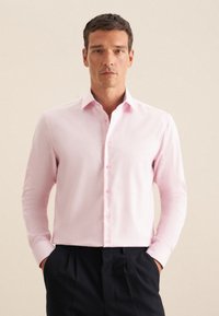 Camicia a maniche lunghe rosa chiaro con colletto classico, bottoni frontali e una texture di tessuto liscia, abbinata a pantaloni scuri.