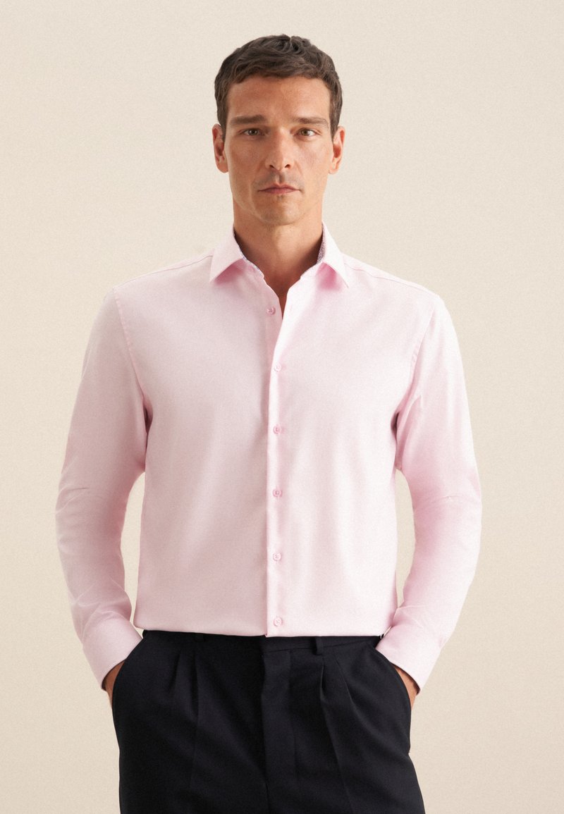 Camicia a maniche lunghe rosa chiaro con colletto classico, bottoni frontali e una texture di tessuto liscia, abbinata a pantaloni scuri.