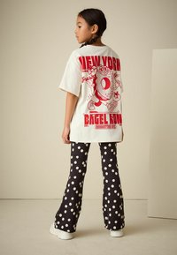T-shirt blanc oversize avec un motif graphique rouge représentant un bagel. Pantalon noir évasé à pois blancs. Des baskets blanches complètent la tenue.