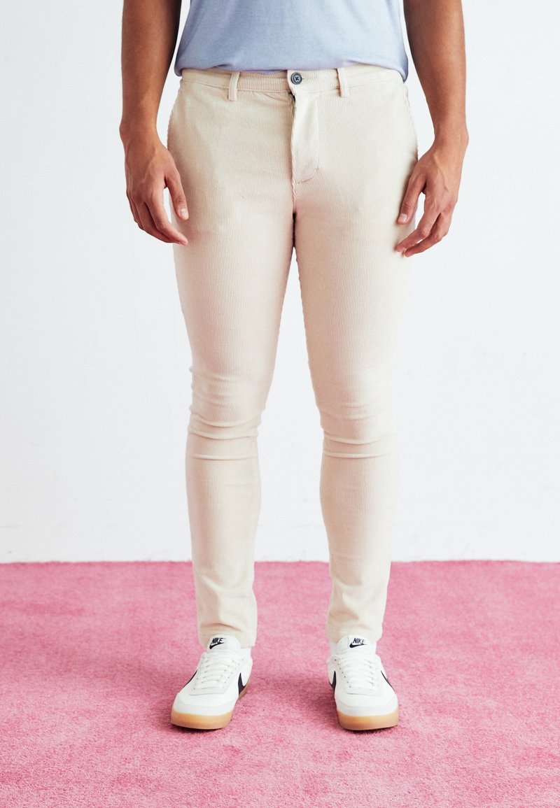 Personne debout sur un tapis rose portant un pantalon en velours côtelé beige, une chemise bleu clair et des baskets blanches Nike avec des semelles en gomme.