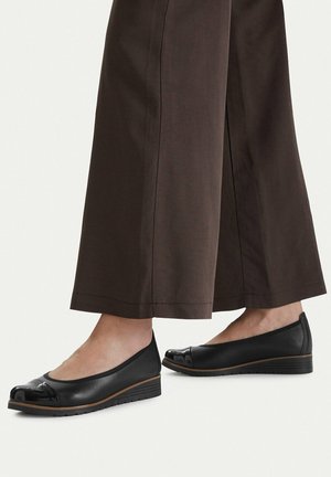 Pies con zapatos planos de cuña de cuero negro con punteras de charol, combinados con pantalones de pierna ancha marrones sobre un fondo liso.