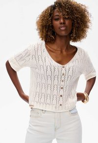 Biała dzianinowa bluzka z krótkim rękawem i dekoltem w serek, z ozdobnym zapięciem na guziki oraz teksturowanym wzorem w zygzak. Noszona na jasnoniebieskie jeansy.
