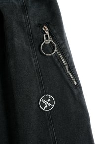 Sección de chaqueta de mezclilla negra con un zipper plateado y un anillo circular, y un parche bordado blanco con detalles en negro.