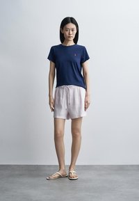 Ženska stoji, oblečena v temno modro majico s kratkimi rokavi, svetlo rožaste kratke hlače z vezalko in bele sandale na enobarvnem sivem ozadju.