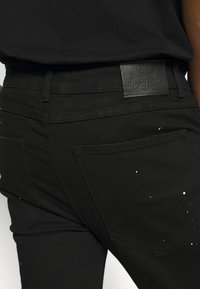 Vaqueros de mezclilla negra con una textura suave, que presentan un parche de cuero y salpicaduras de pintura blanca cerca de los bolsillos traseros.