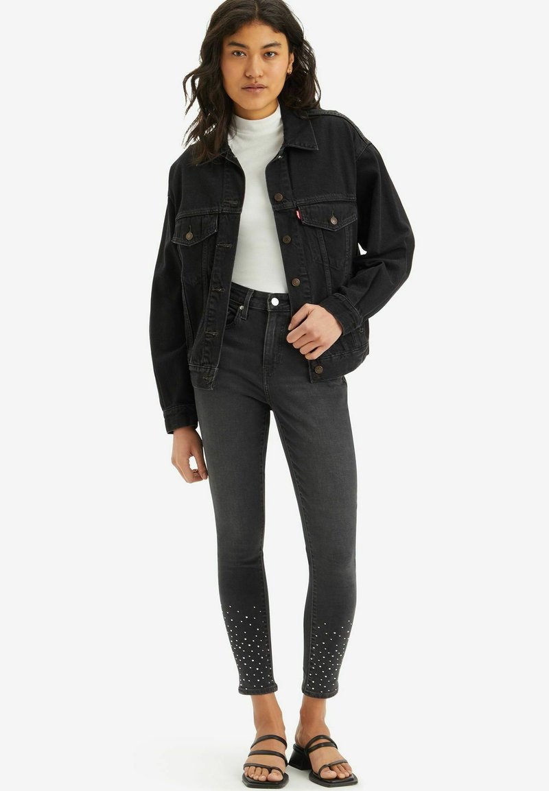 Schwarze Jeansjacke und dunkle Skinny-Jeans mit silbernen Verzierungen an den Bündchen, kombiniert mit einem weißen Rollkragenpullover und schwarzen Sandalen.