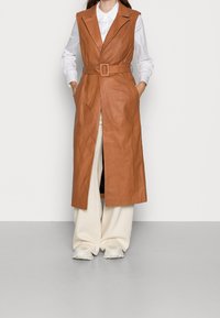 Chaleco sin mangas de cuero marrón con cintura cinturón, que presenta un cuello con muesca y bolsillos laterales, combinado con pantalones de pierna ancha color crema.